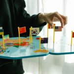 Mão organizando bandeiras de diferentes países sobre um mapa-múndi.