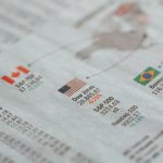 Close de um jornal com estatísticas de mercados financeiros globais e bandeiras de diferentes países.