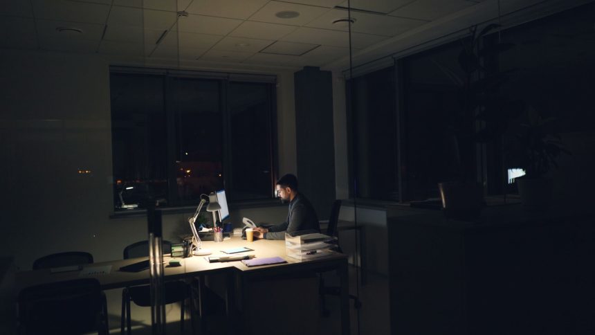 Homem trabalhando sozinho à noite em um escritório, iluminado pela luz da mesa.